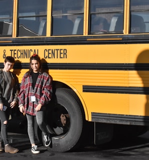 Two people are standing in front of a yellow school bus with the text 'County Career & Technical Center' written on its side. Both individuals are wearing casual clothing and are posed next to the bus shadow on the ground.