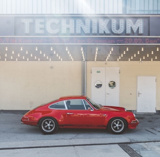 A vintage red sports car is parked in front of a building with a sign reading 'TECHNIKUM'. The structure has a retro design with a row of round light bulbs beneath the sign. There are three doors on the front of the building, with a neutral tone wall.