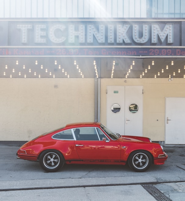 A vintage red sports car is parked in front of a building with a sign reading 'TECHNIKUM'. The structure has a retro design with a row of round light bulbs beneath the sign. There are three doors on the front of the building, with a neutral tone wall.