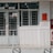 A building entrance with metal barred doors and windows, featuring signs indicating it belongs to a faculty for high-quality training. The facade is white, and there are red fire extinguishers and safety notices mounted on the wall next to the entrance.