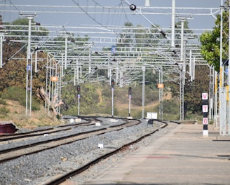 A railway station's tracks extend into the distance under a network of overhead electrical wires. Signal lights and signs are visible along the tracks, with trees and vegetation alongside. A platform runs parallel to the tracks, with a sign reading 'VTO' on it.