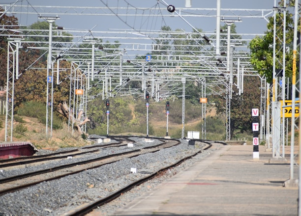 A railway station's tracks extend into the distance under a network of overhead electrical wires. Signal lights and signs are visible along the tracks, with trees and vegetation alongside. A platform runs parallel to the tracks, with a sign reading 'VTO' on it.