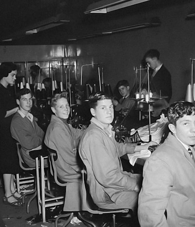 A group of young men and one woman are seated in a sewing workshop, with sewing machines and spools of thread visible. The men are wearing work attire and are seated in front of machines, seemingly involved in textile work. The setting appears to be a vocational or educational environment.