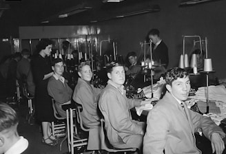 A group of young men and one woman are seated in a sewing workshop, with sewing machines and spools of thread visible. The men are wearing work attire and are seated in front of machines, seemingly involved in textile work. The setting appears to be a vocational or educational environment.