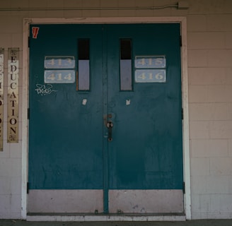 A set of teal double doors with room numbers 413 to 416 displayed. The doors are situated in a brick wall with light gray paint, showing signs of aging and some graffiti. On the left side, vertical signs read 'Technology' and 'Education'. The environment hints at an institutional or school setting.