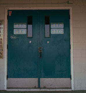 A set of teal double doors with room numbers 413 to 416 displayed. The doors are situated in a brick wall with light gray paint, showing signs of aging and some graffiti. On the left side, vertical signs read 'Technology' and 'Education'. The environment hints at an institutional or school setting.