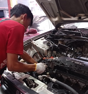 A mechanic is working on an open car engine, with visible components including the Honda VTEC engine. The person is wearing a red shirt and white gloves, focused on adjusting or repairing the engine. There are several cars in the background, indicating a workshop environment.