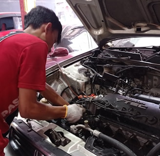 A mechanic is working on an open car engine, with visible components including the Honda VTEC engine. The person is wearing a red shirt and white gloves, focused on adjusting or repairing the engine. There are several cars in the background, indicating a workshop environment.