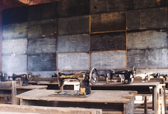 A classroom setting features several old-fashioned sewing machines on wooden desks lined in rows. The background includes a wall of chalkboards covered with writing, creating an atmosphere of a vintage educational or vocational training environment.