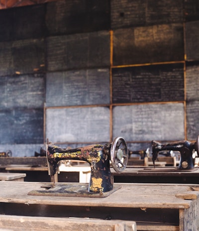 A classroom setting features several old-fashioned sewing machines on wooden desks lined in rows. The background includes a wall of chalkboards covered with writing, creating an atmosphere of a vintage educational or vocational training environment.