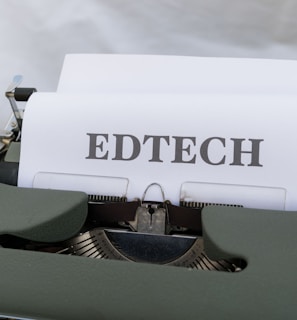 A vintage typewriter with a sheet of paper that has the word 'EDTECH' typed on it. The typewriter casing is dark green and the setting appears to be on a light wooden surface.